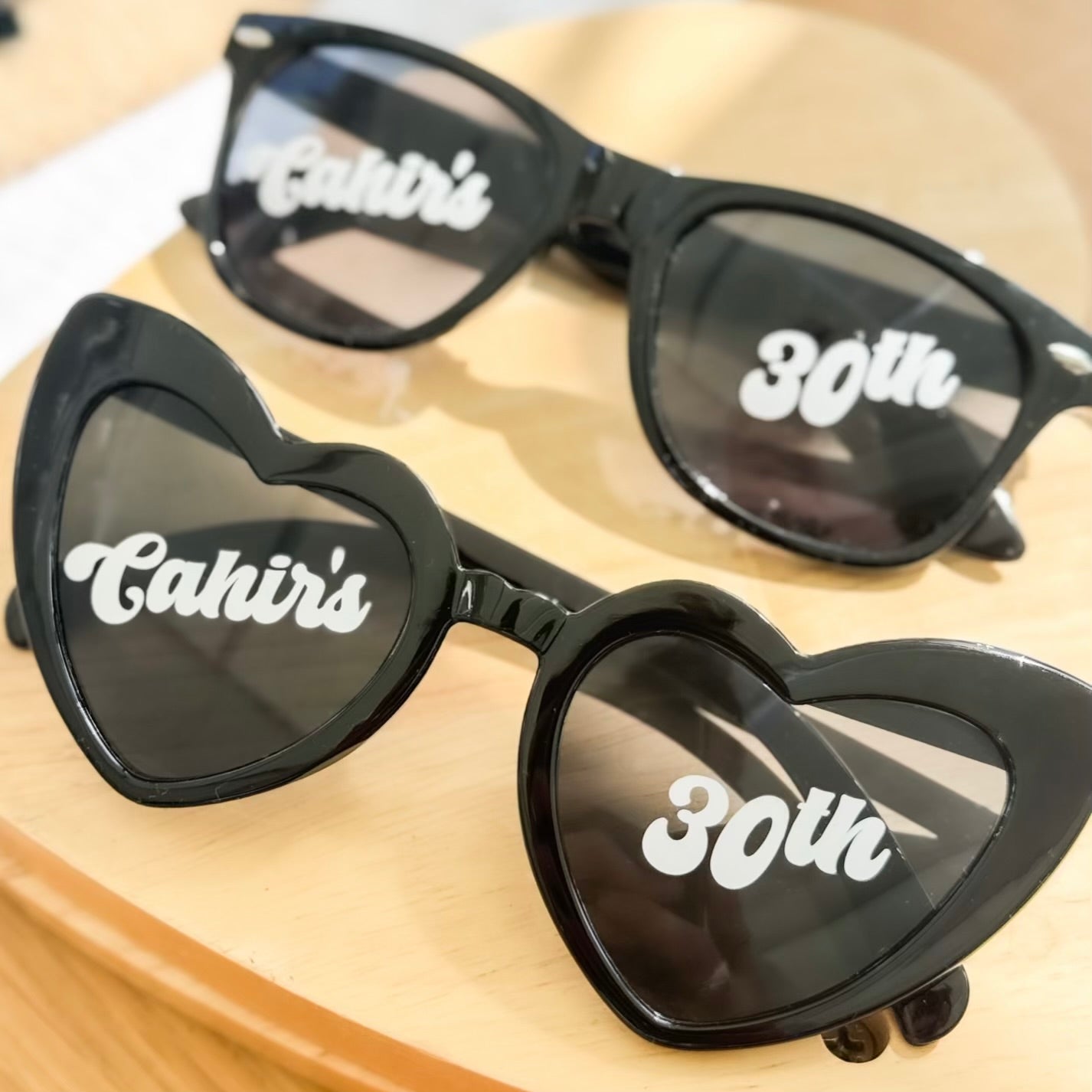 Heart-shaped black sunglasses with ‘Cahir’s’ and ‘30th’ text on wooden surface