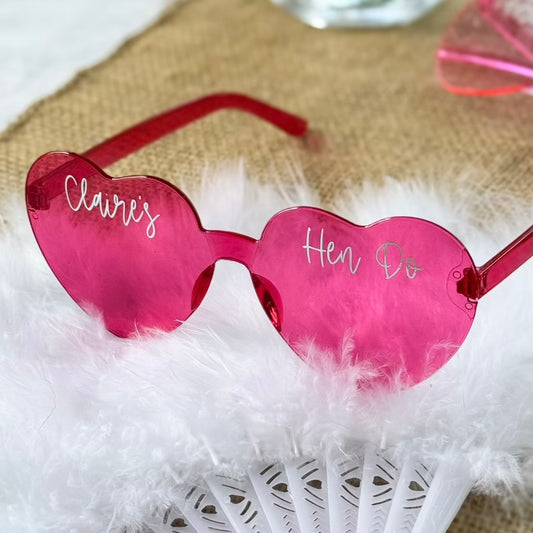 Pink heart-shaped sunglasses with 'Claire's Hen Do' text on a fluffy white surface.