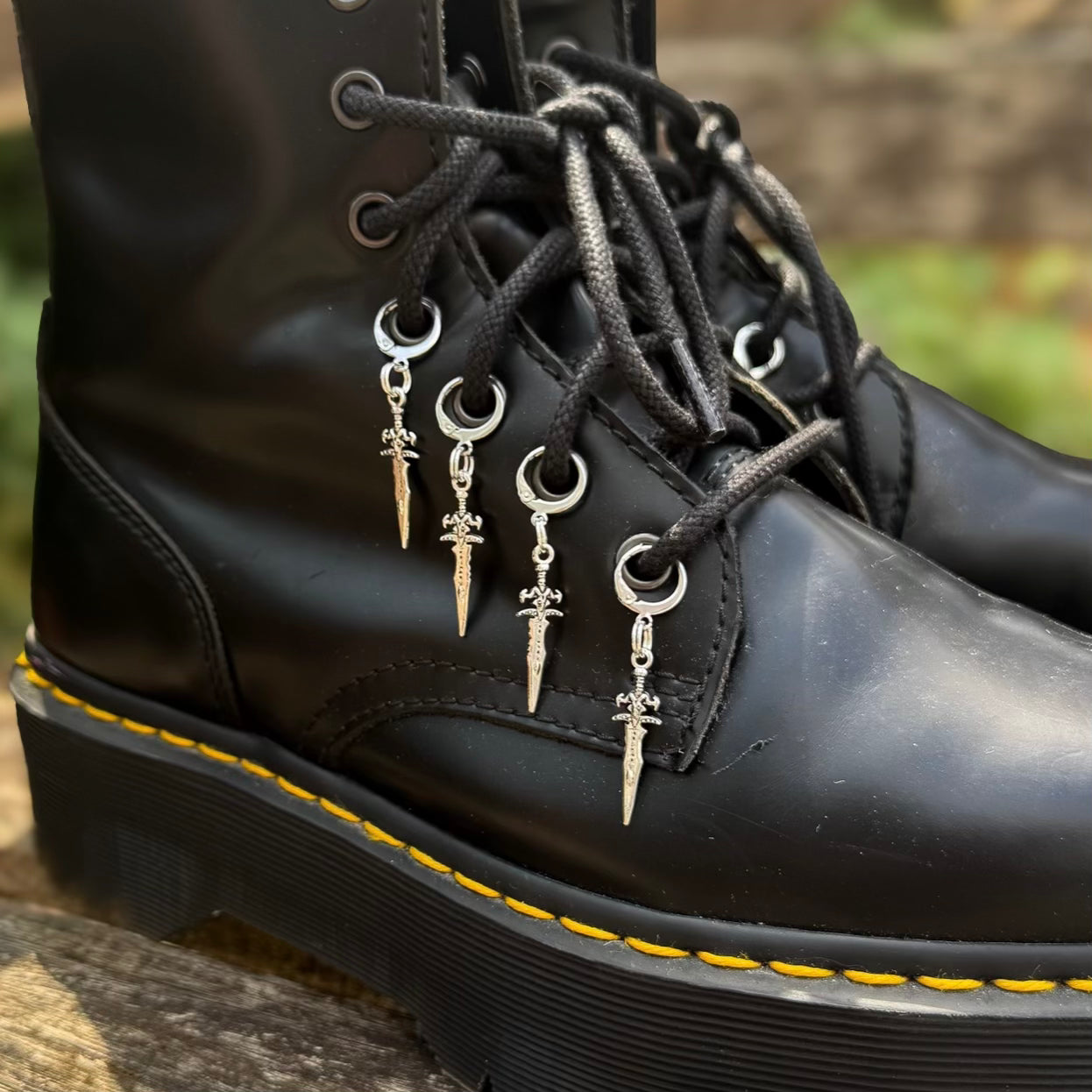 A close-up of a black leather lace-up boot with silver metal eyelets and dangling dagger charms threaded through the laces. The boot has a black sole with yellow stitching, resting outdoors on a wooden surface with a blurred natural background.