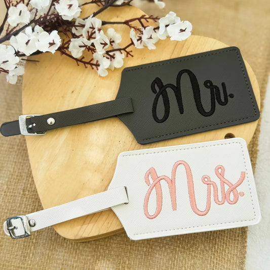 Black 'Mr.' and white 'Mrs.' luggage tags on a wooden surface with flowers.
