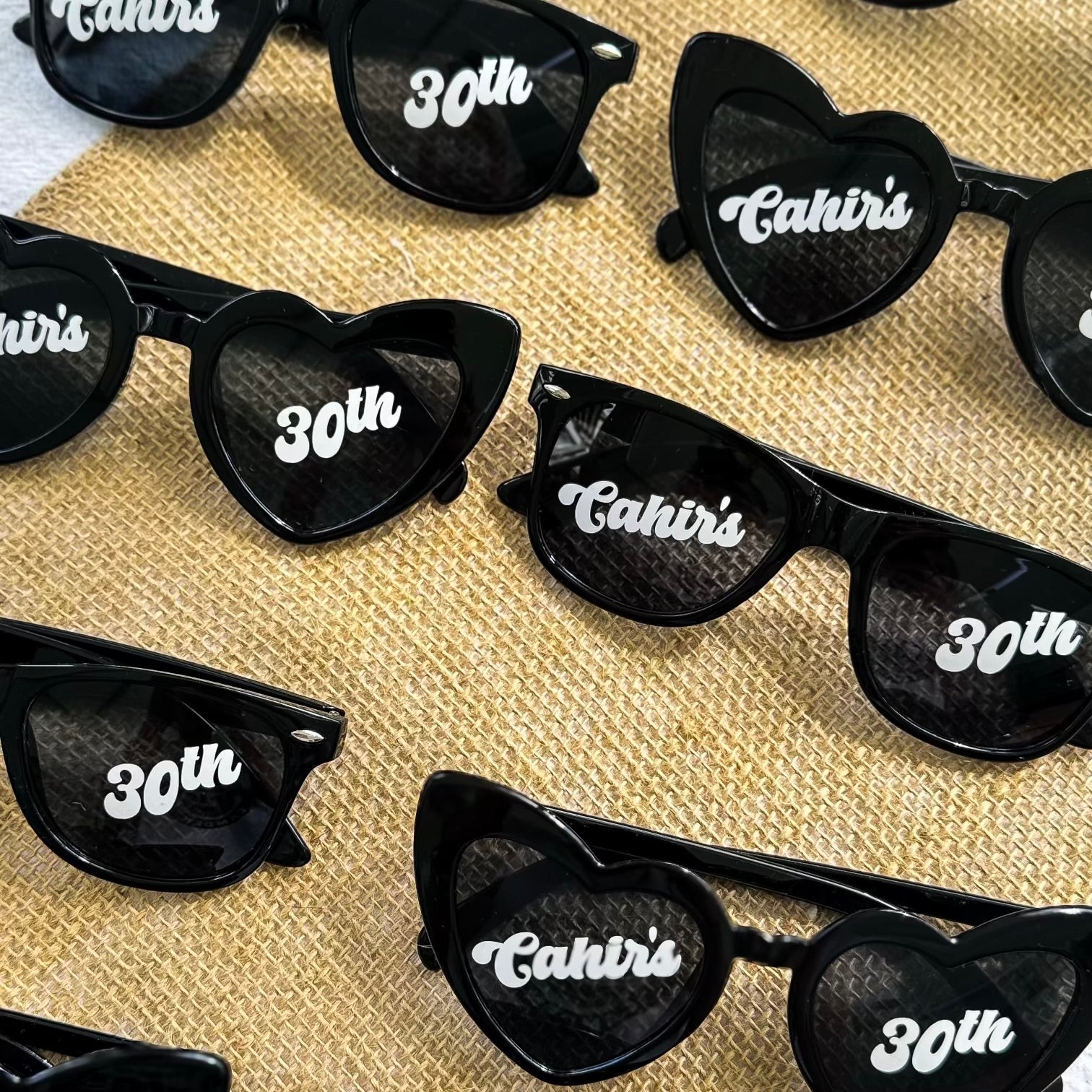 Heart-shaped black sunglasses with 'Cahir's 30th' text on a textured surface