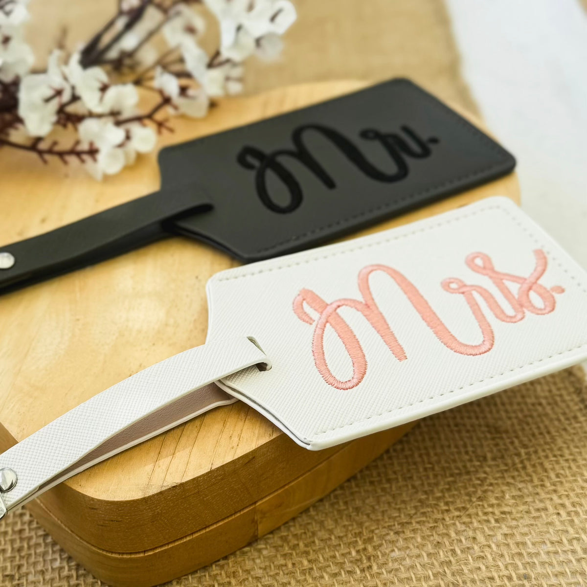Two luggage tags, one black with 'Mr.' and one white with 'Mrs.', on a wooden surface.