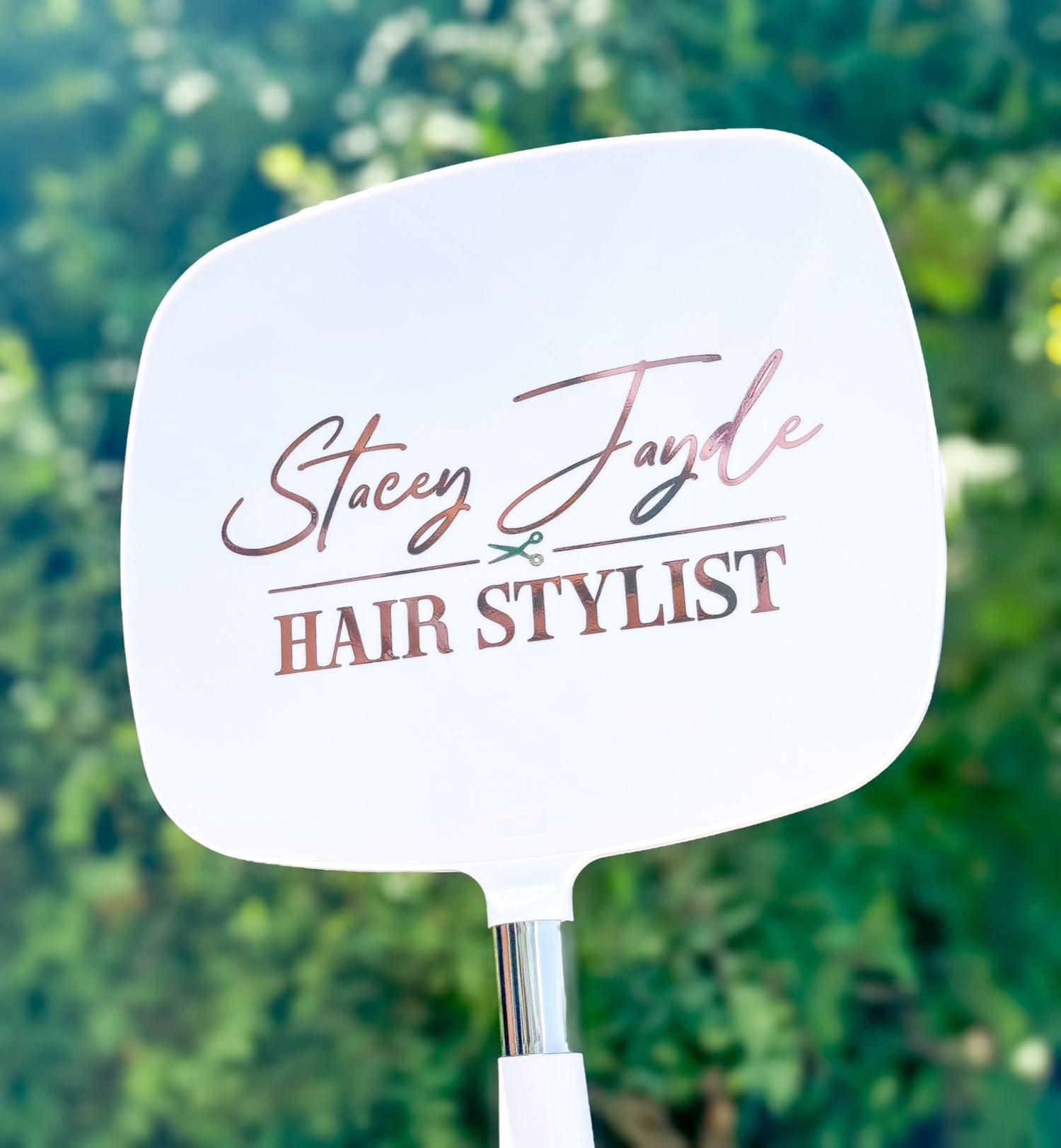 Handheld mirror with 'Stacey Jayde Hair Stylist' branding against a blurred green background