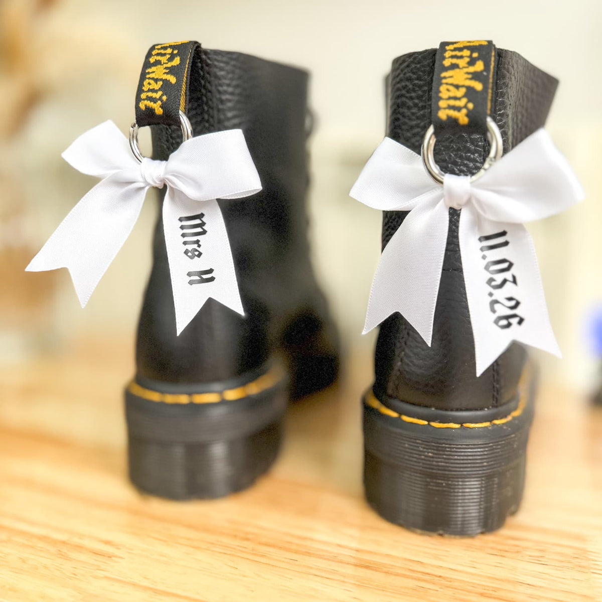 A pair of black Doc Marten boots with white satin bows attached to them, displaying personalized text that reads 'Mrs H' and a date '11.05.