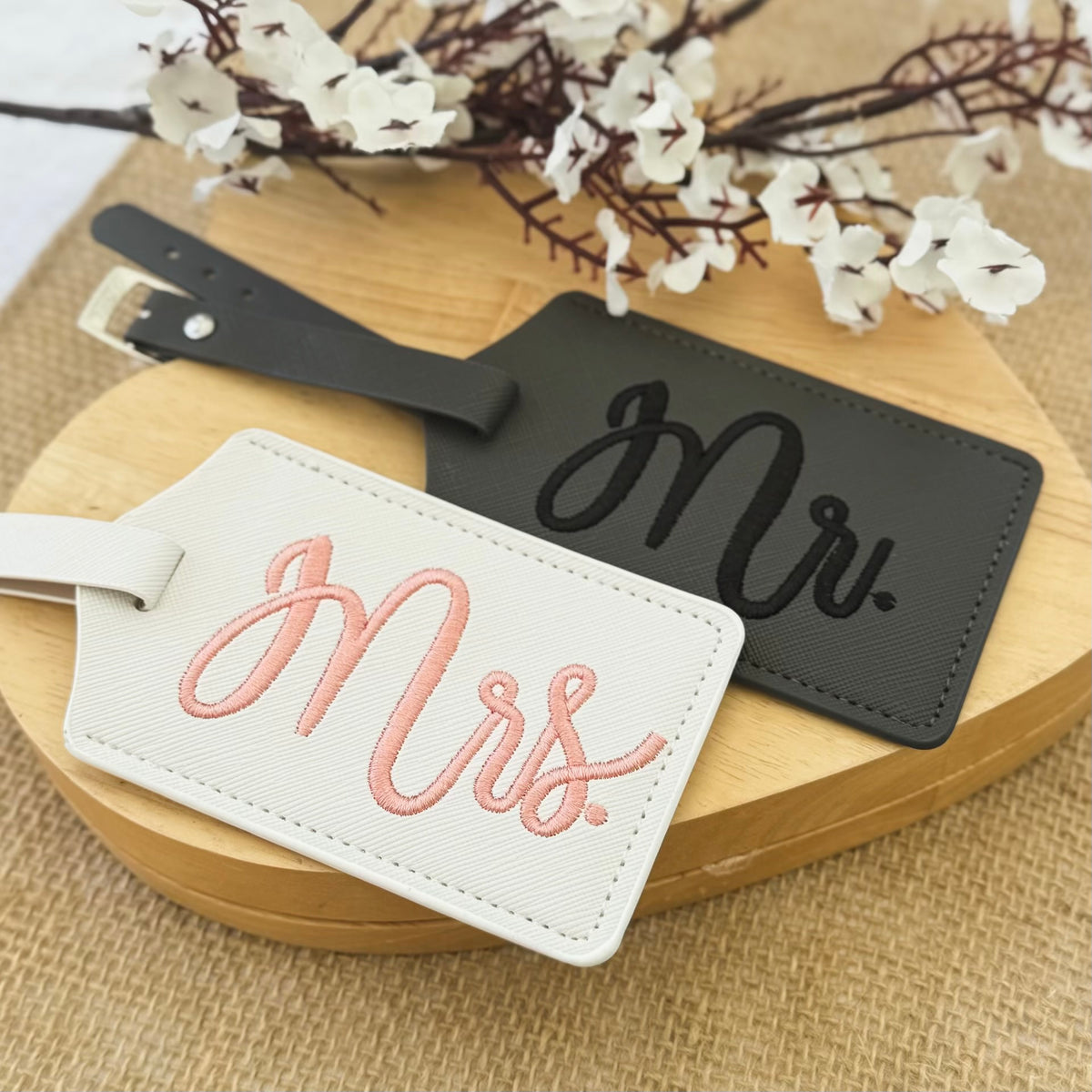 Two luggage tags, one white with 'Mrs.' and one black with 'Mr.', on a wooden surface with flowers.