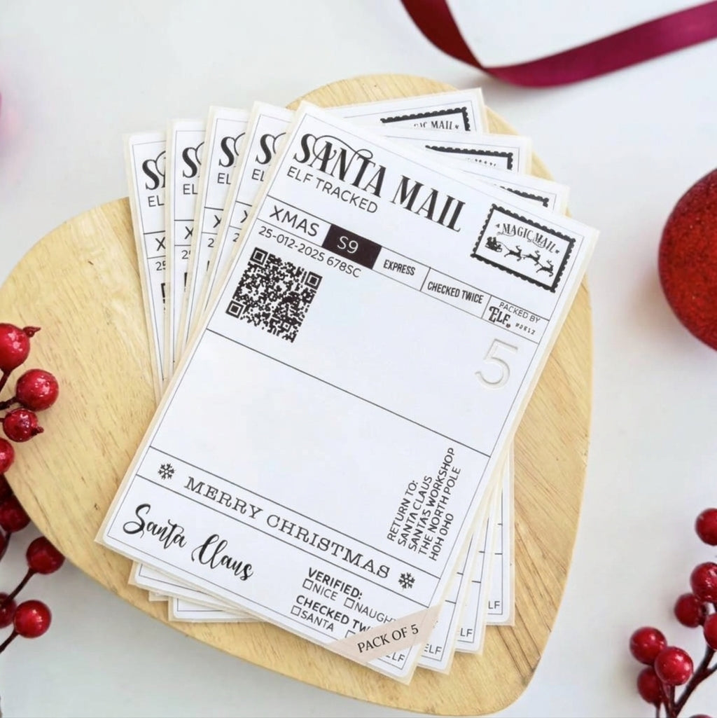 A stack of Santa-themed Christmas elf tracking mailer envelopes displayed on a round wooden board, surrounded by festive red berries and a red ribbon.