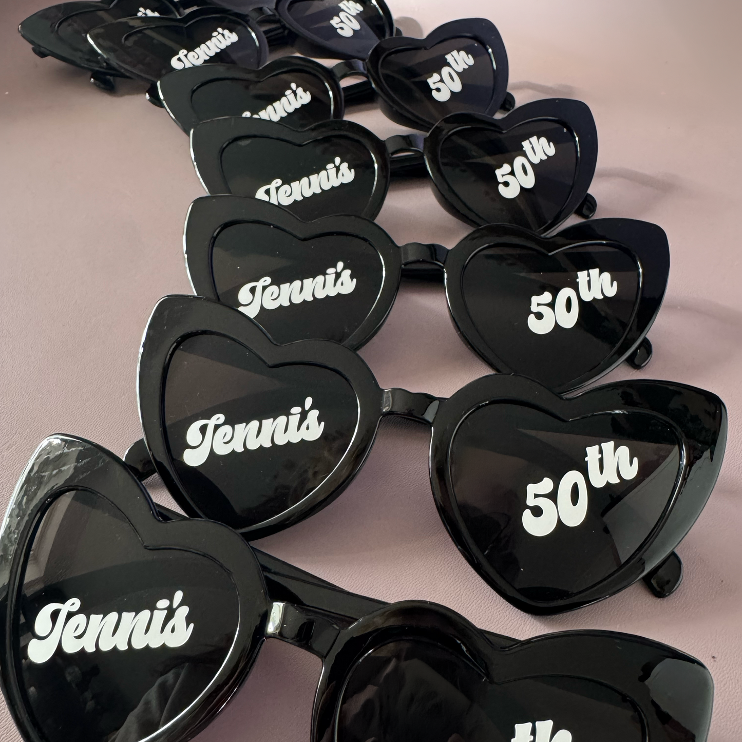Heart-shaped sunglasses with 'Jenni's 50th' text on a beige background