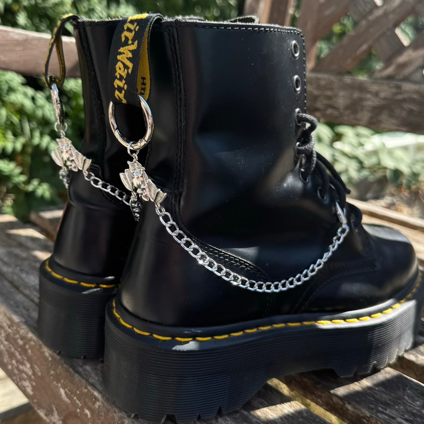 A pair of black leather platform combat boots with silver chain leg straps and dangling charms, resting on a wooden bench outdoors.
