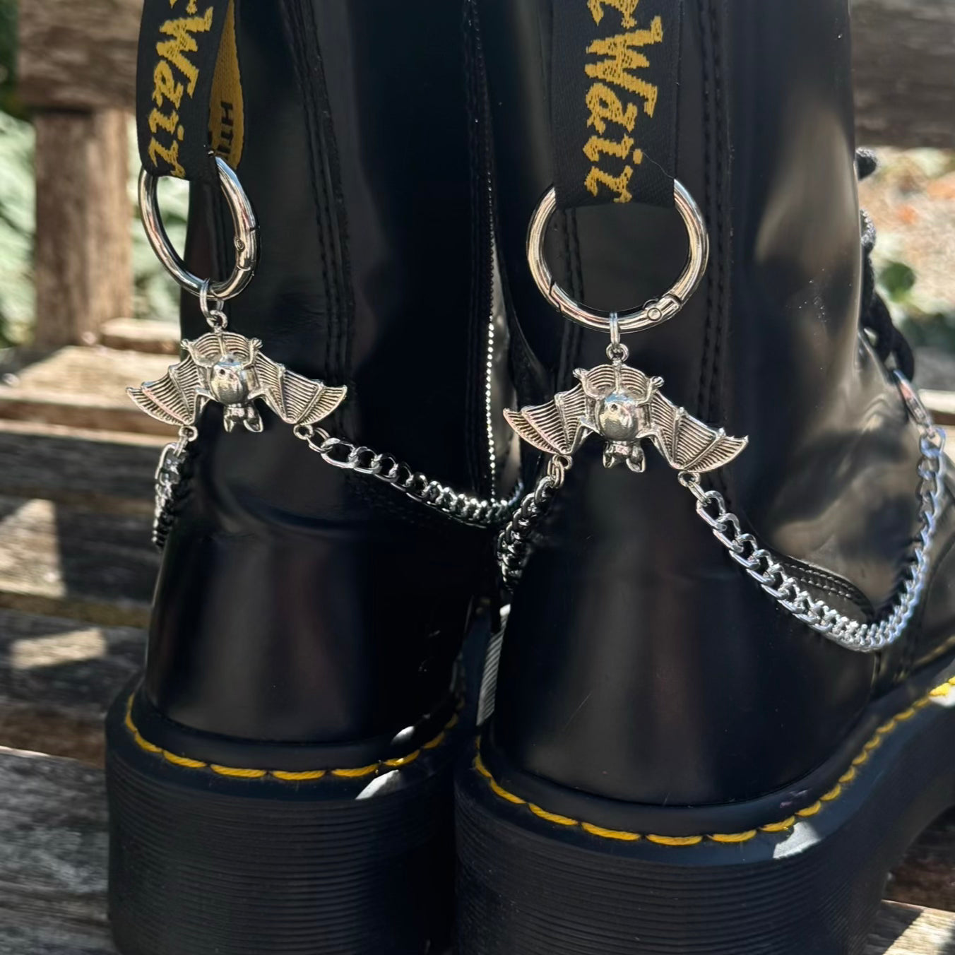 Close-up of a pair of black leather boots with silver winged crescent moon chain accessories attached near the back, shown outdoors on a wooden surface.