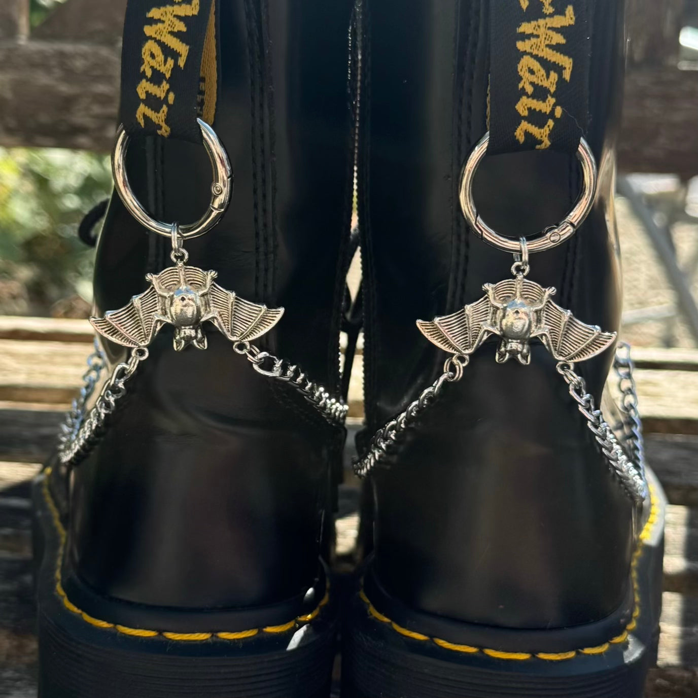 Close-up of the back of a pair of black leather boots with silver bat-shaped keychains attached by chains, shown outdoors on a wooden surface.
