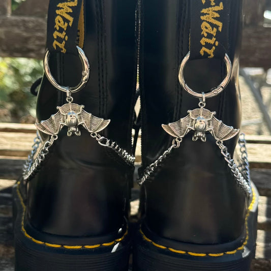 Close-up of the back of a pair of black leather boots with silver bat-shaped keychains attached by chains, shown outdoors on a wooden surface.