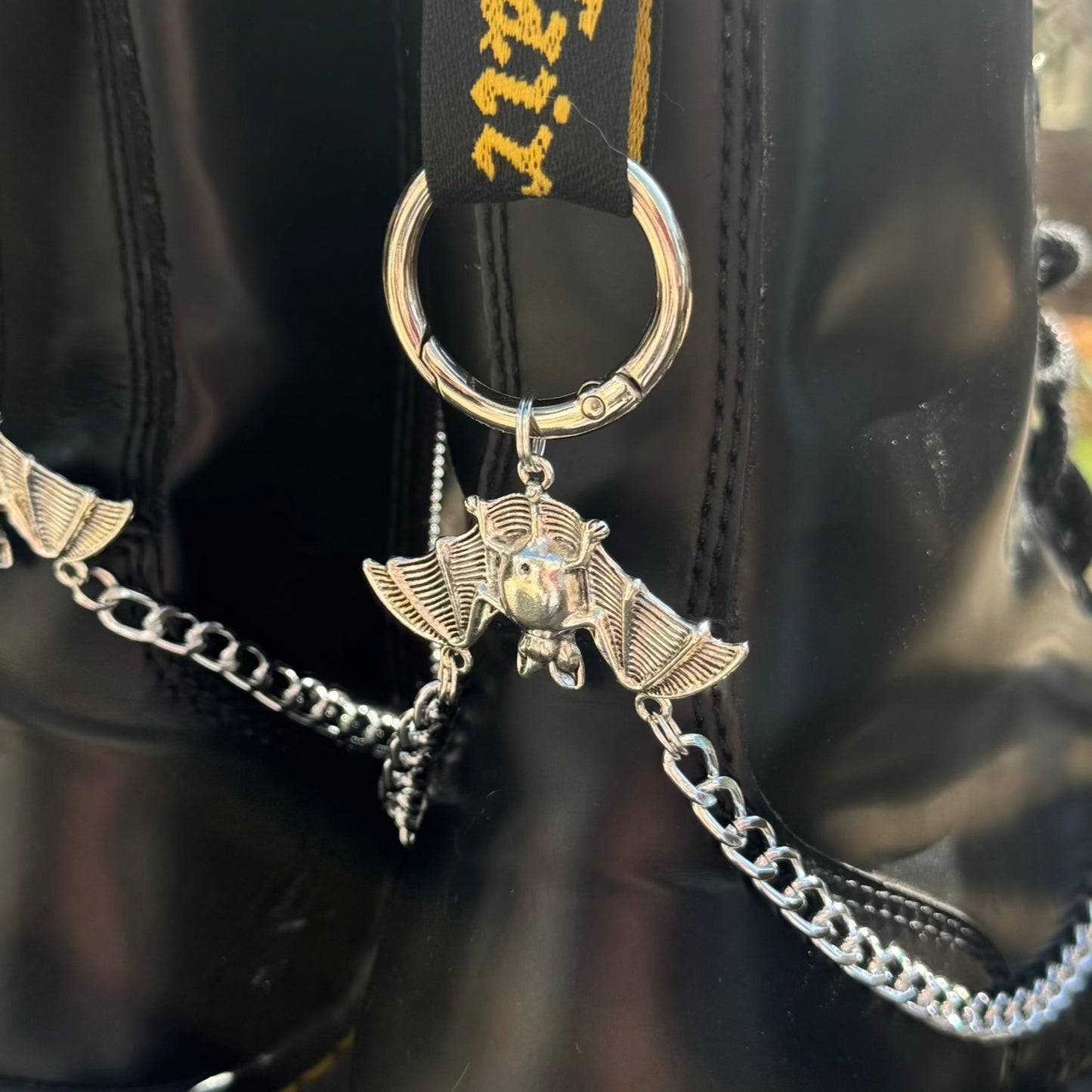 A silver metal keychain with a bat charm and chain, attached to a black strap with gold lettering, shown against a black leather background.