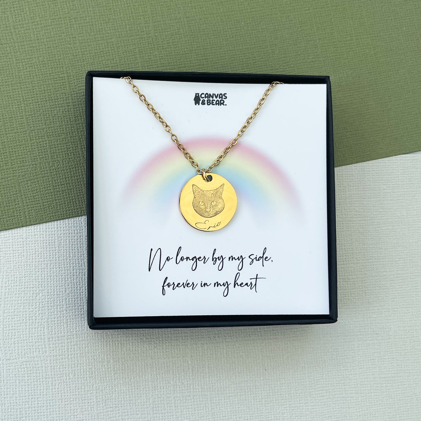 An example of a personalised necklace. A gold necklace with a cat engraved with the name Eric  below. This image features a gift bow with a gift card reading "no longer by my side, forever in my heart' with a light rainbow above