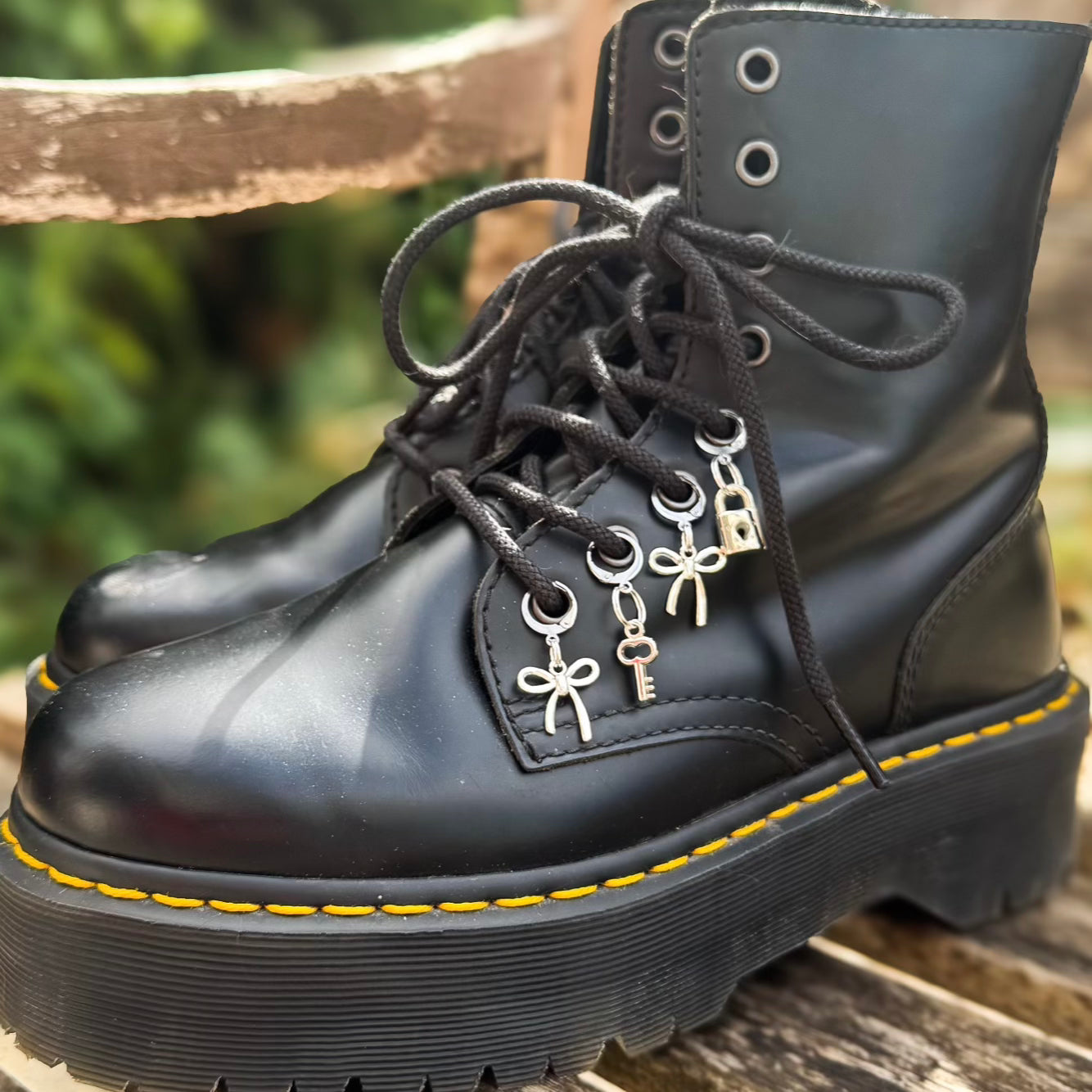 A pair of black leather lace-up combat boots with silver bow, key, and padlock charms threaded through the eyelets, resting on a wooden surface with a blurred outdoor background.
