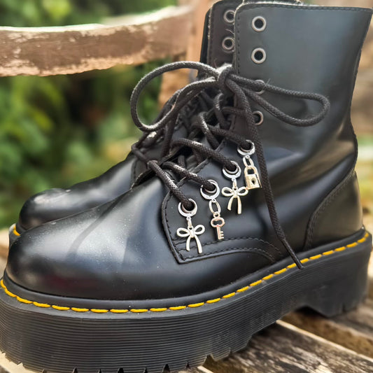 A pair of black leather lace-up combat boots with silver bow, key, and padlock charms threaded through the eyelets, resting on a wooden surface with a blurred outdoor background.