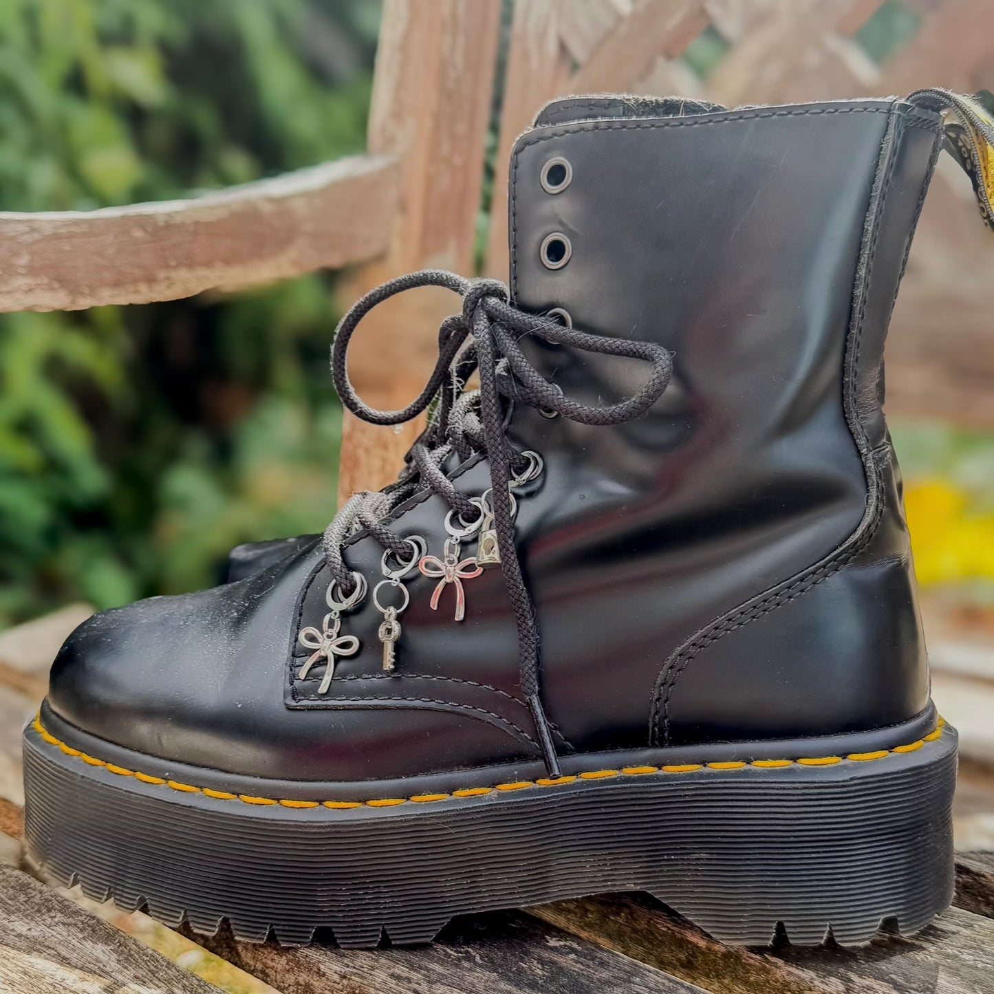 A black leather combat boot with a chunky sole, shown on a wooden surface with a blurred outdoor background. The boot features decorative silver metal charms on the laces.