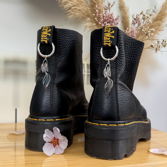 Pair of black leather Dr. Martens boots featuring silver metallic Lucky Koi Fish boot charms hanging from the heel loops, styled on a wooden table with a cherry blossom flower.