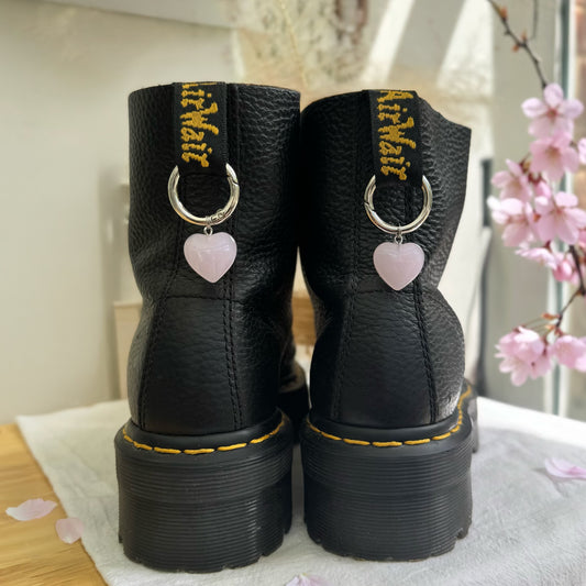Black boots with heart-shaped charms on a light surface with pink flowers in the background