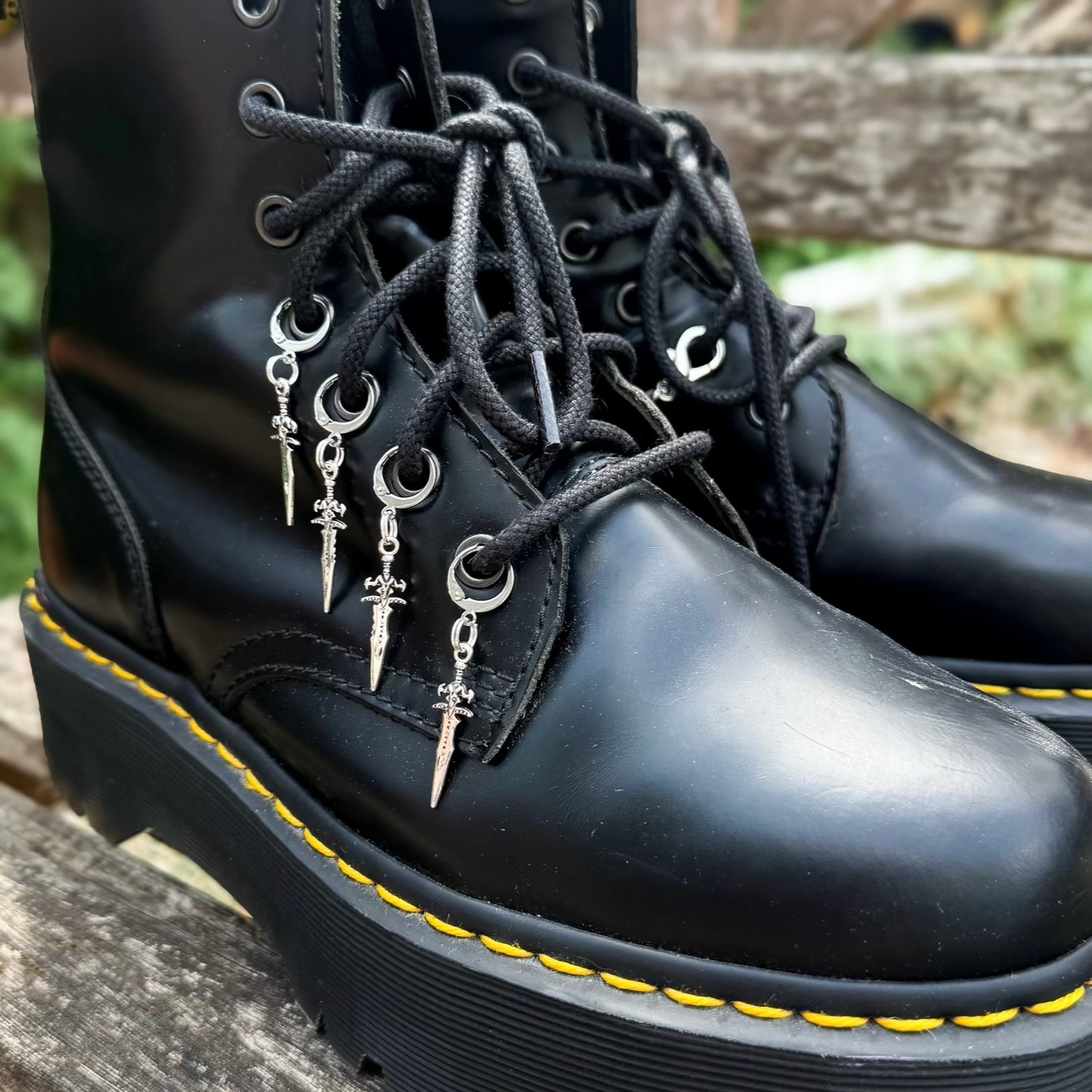 A close-up of a black leather lace-up boot with silver metal eyelets and dangling silver charms shaped like swords charms
